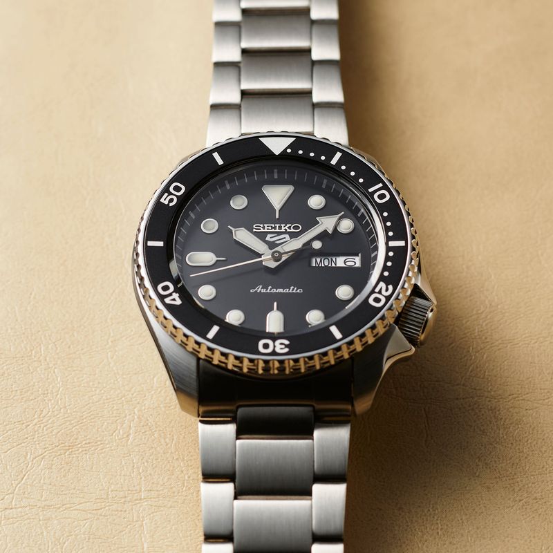 Relógio de pulso Seiko com mostrador preto, vidro espesso, pulseira de metal prateada e detalhes em dourado na moldura.