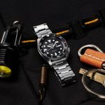 Relógio de mergulho Seiko com pulseira de metal, rodeado por equipamentos de escalada em fundo preto.