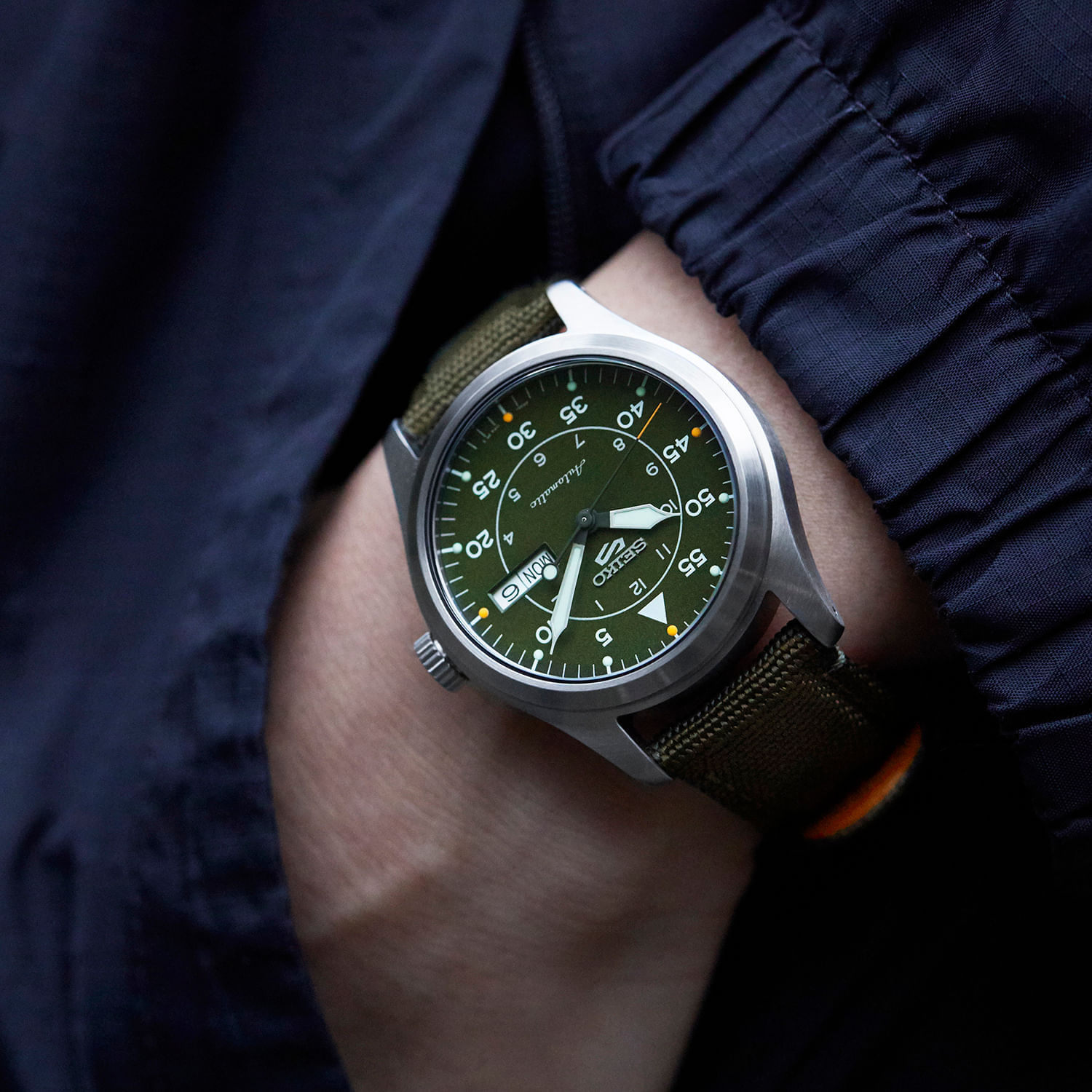 Relógio com mostrador verde, branco e amarelo, com várias funções e marcas, preso a um pulso usando uma pulseira de tecido verde e uma jaqueta escura.