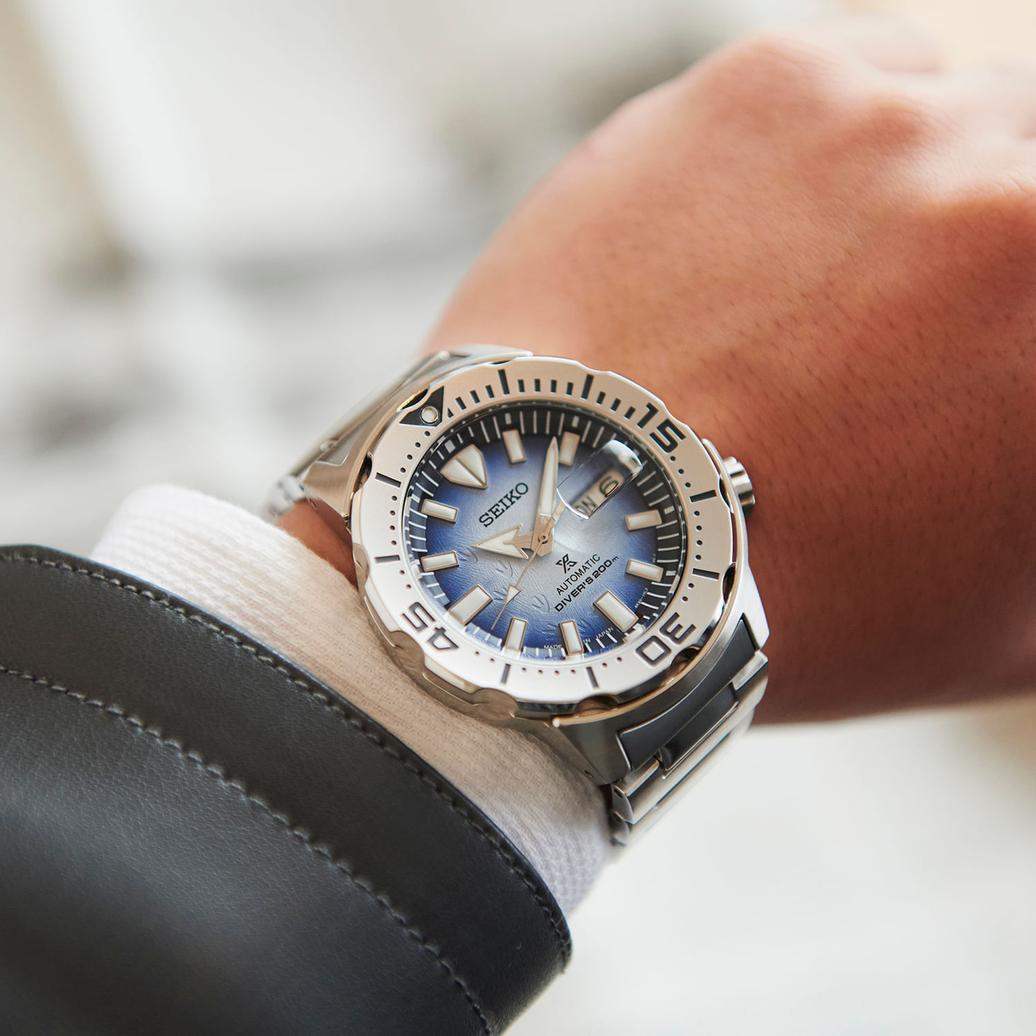 Relógio de pulso com pulseira de metal e caixa prateada, mostrador azul com índices e numbers marcadores, em um pulso com roupa social preta e branca.