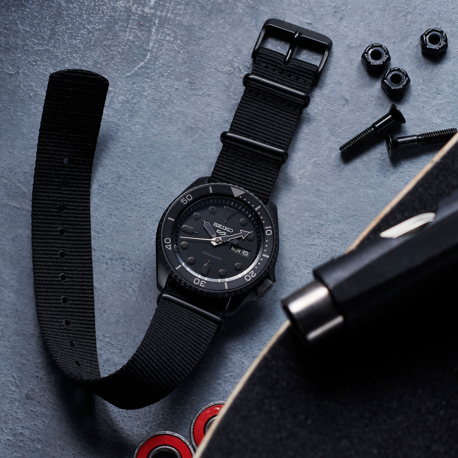 Relógio preto com pulseira de tecido, mostrador analógico com dia e data, junto a skate, parafusos e rolamentos.