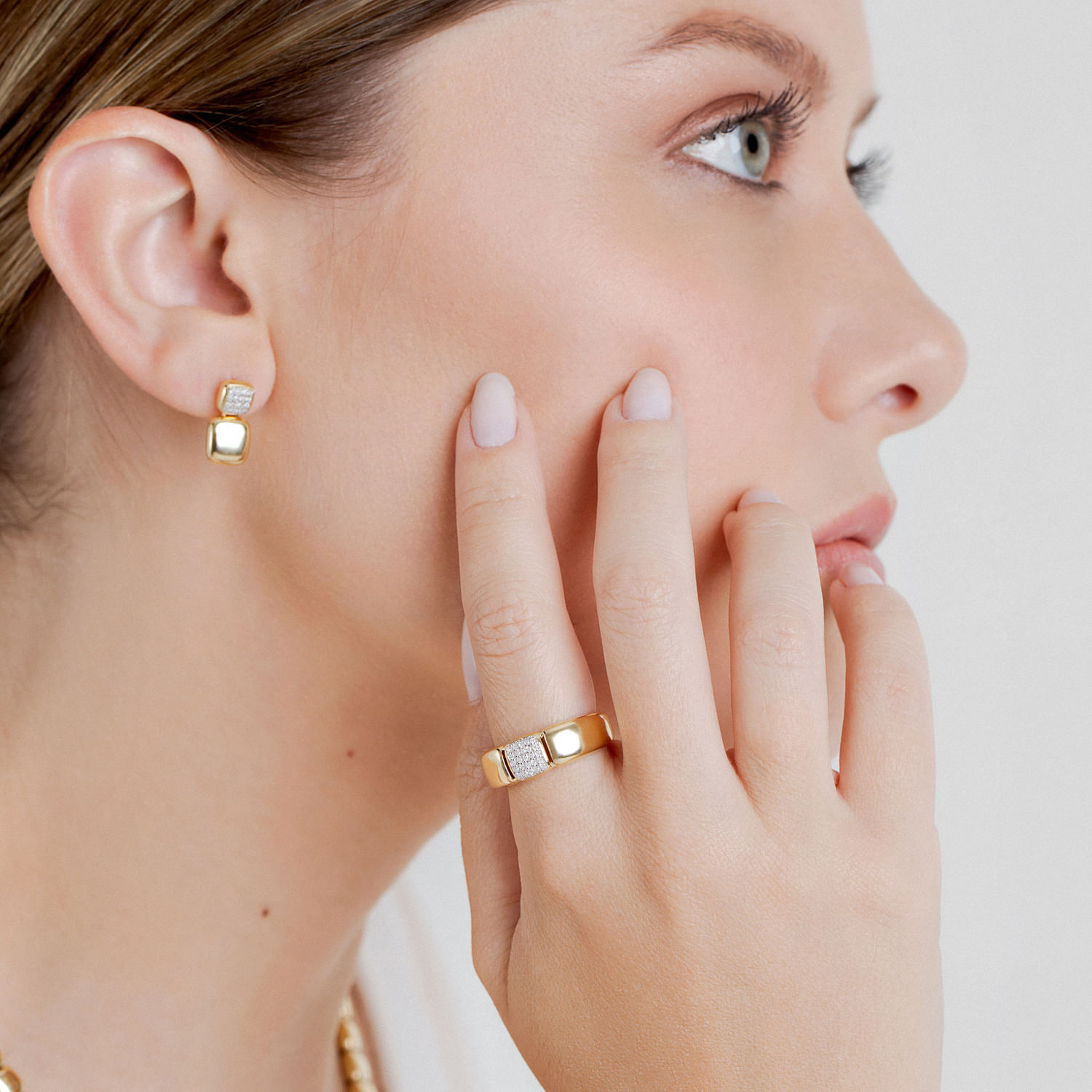 Close de uma mulher usando joias de ouro e diamantes, incluindo brincos, um anel e um colar, com a mão perto do rosto.