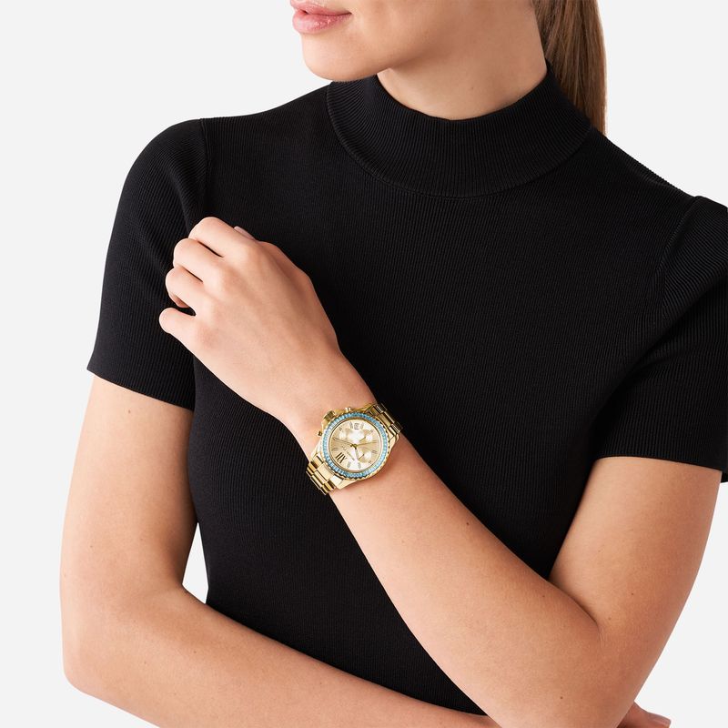 Muher com relógio dourado e pulseira na mão esquerda, vestindo blusa preta de manga curta e gola alta.