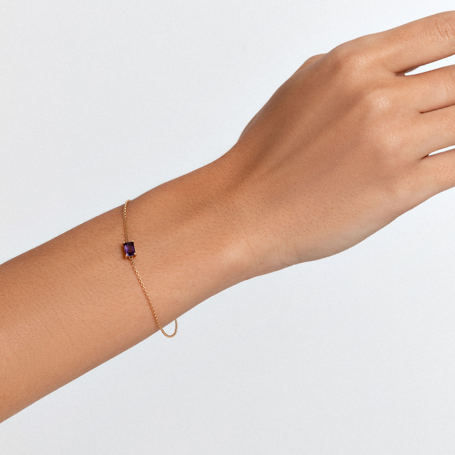 Pulseira com corrente fina e pedra rectangular roxa no centro, usada no pulso de uma mão.