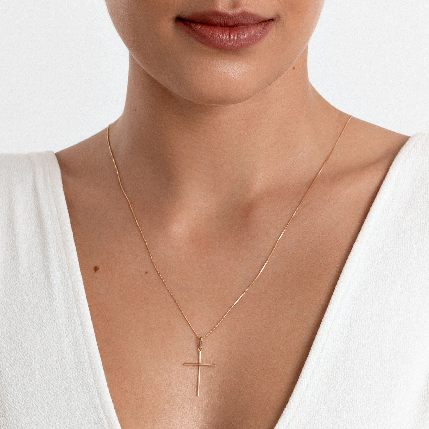 Colar delicado de corrente fina com pingente em forma de cruz dourado, usado por uma mulher com roupa branca.