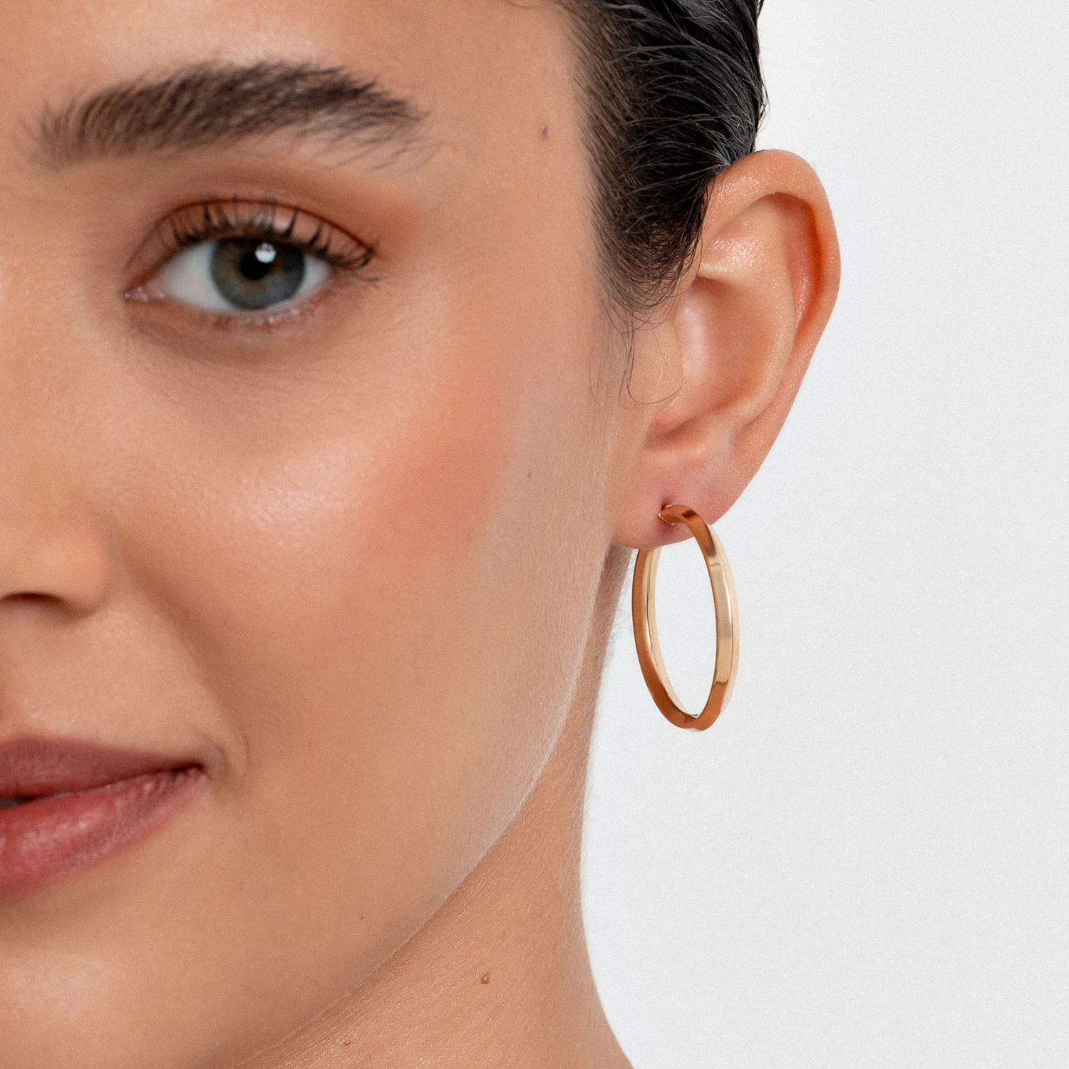 Close-up de uma mulher com brinco de argola dourado na orelha direita, pele clara, sobrancelhas arqueadas e olhos azuis.