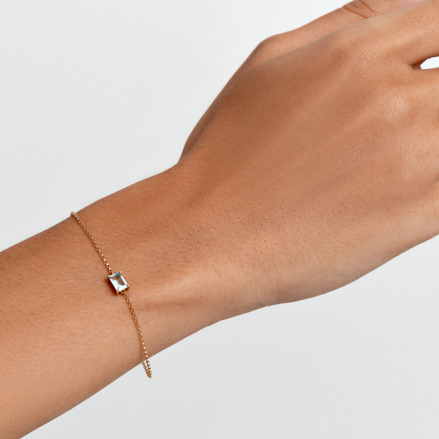 Pulseira dourada com pingente de pedra rectangular translúcida na pele de uma mão.