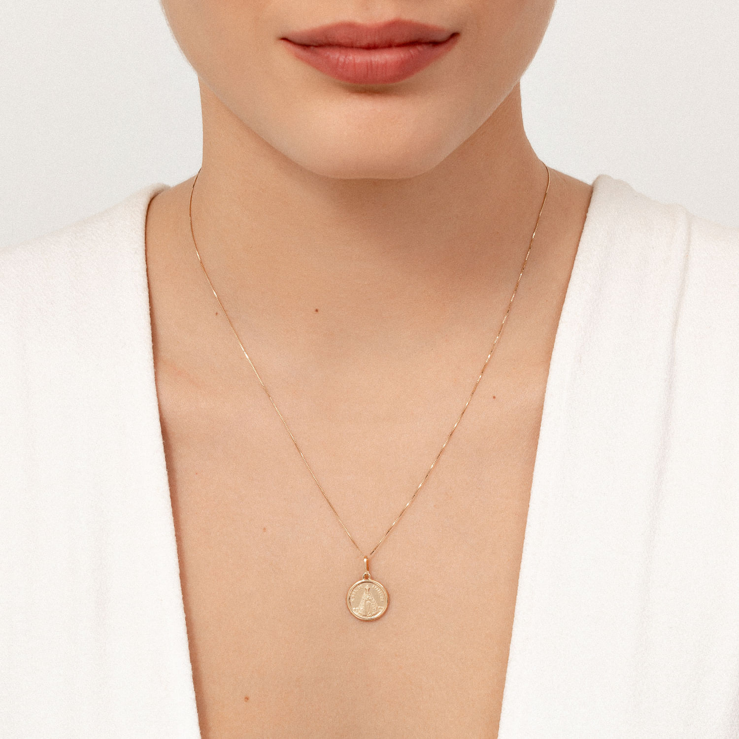 Gargantilha delicada de corrente fina com pingente circular dourado, usado no pescoço de uma mulher com pele clara e blusa branca.