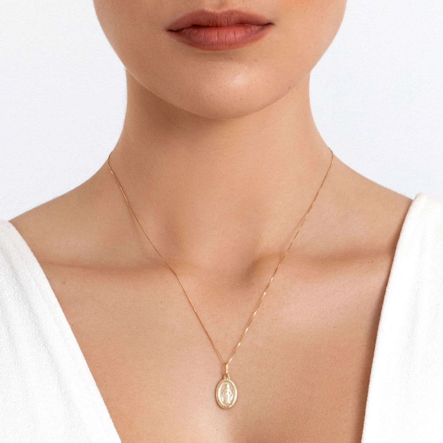 Colar dourado delicado com medalha oval com imagem de uma santa, usado por uma mulher com vestido branco.