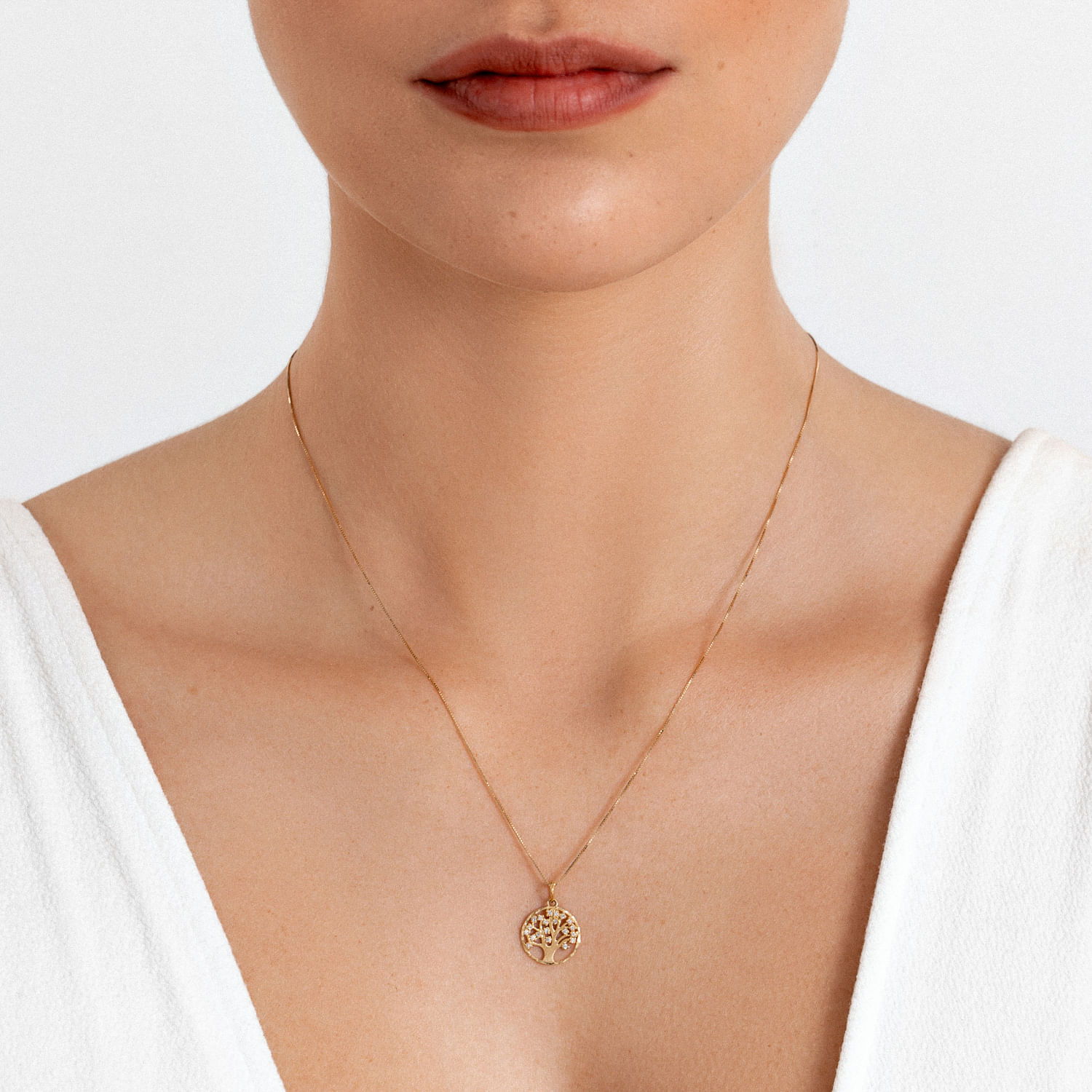 Colar dourado com pingente de árvore da vida, usado por uma mulher com blusa branca.