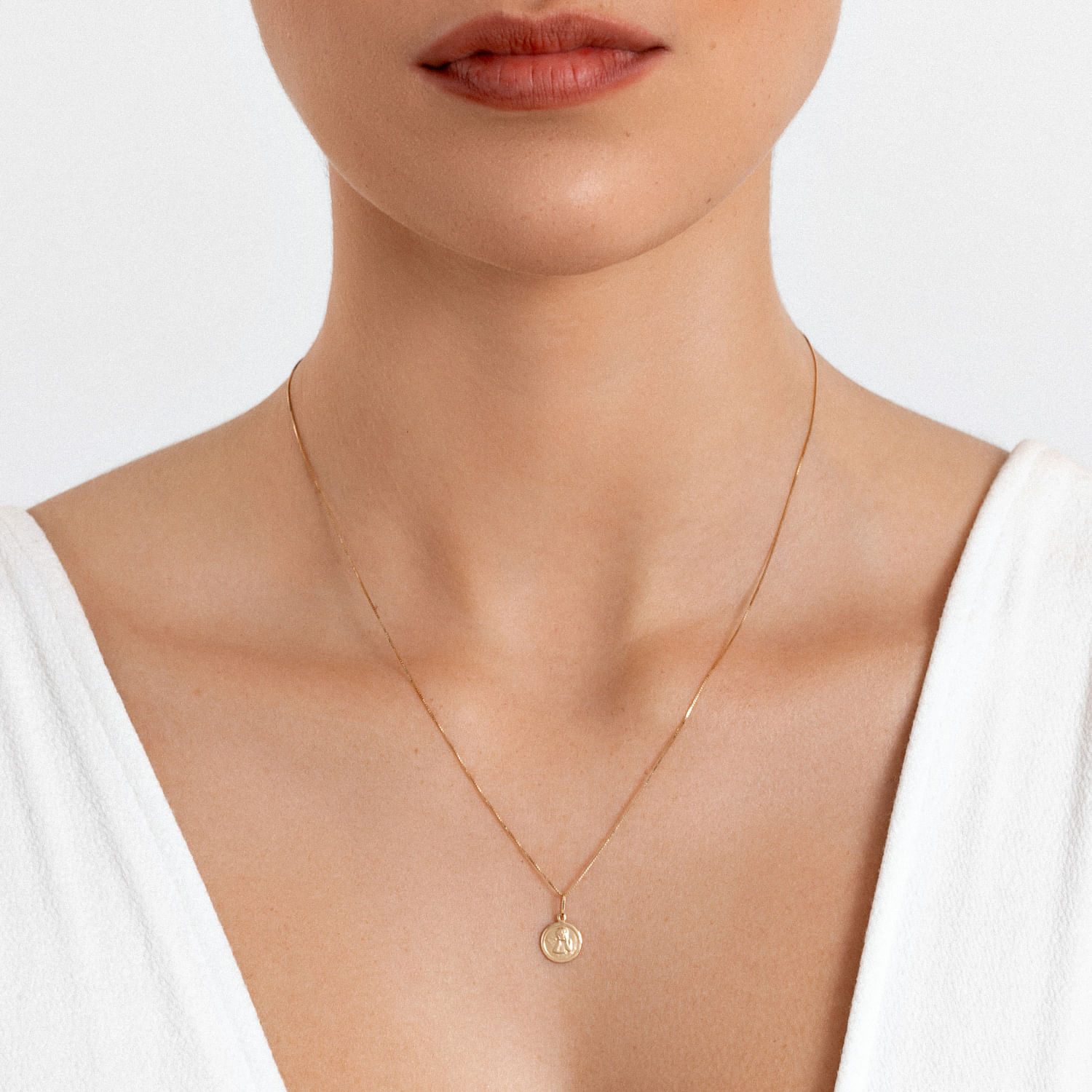 Colar dourado delicado com pingente circular e letra "A" na frente, usado por uma mulher com blusa branca.