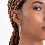 Close-up da orelha de uma mulher com cabelo trançado, usando um brincinho dourado com design ondulado.