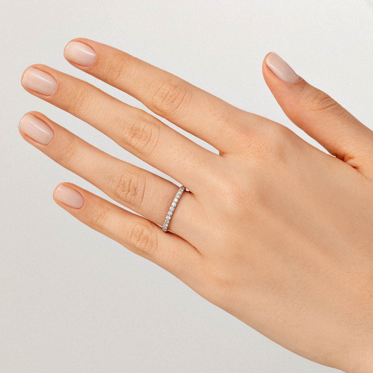 Mão com unhas pintadas de nude, usando anel com pedras brilhantes na dedo médio.