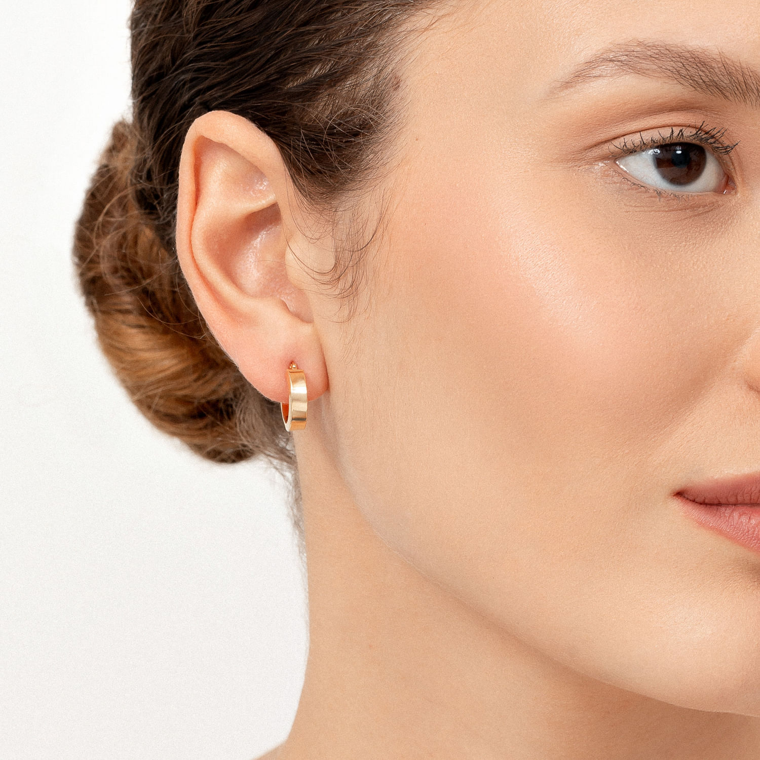 Close-up de uma mulher usando um brinco dourado pequeno e redondo na orelha esquerda, com pele clara e cabelo castanho preso atrás.