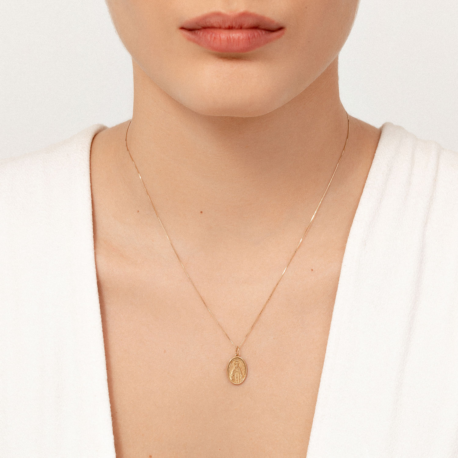 Colar dourado com pingente oval decorado, usado por uma mulher com pele clara e camisa branca.