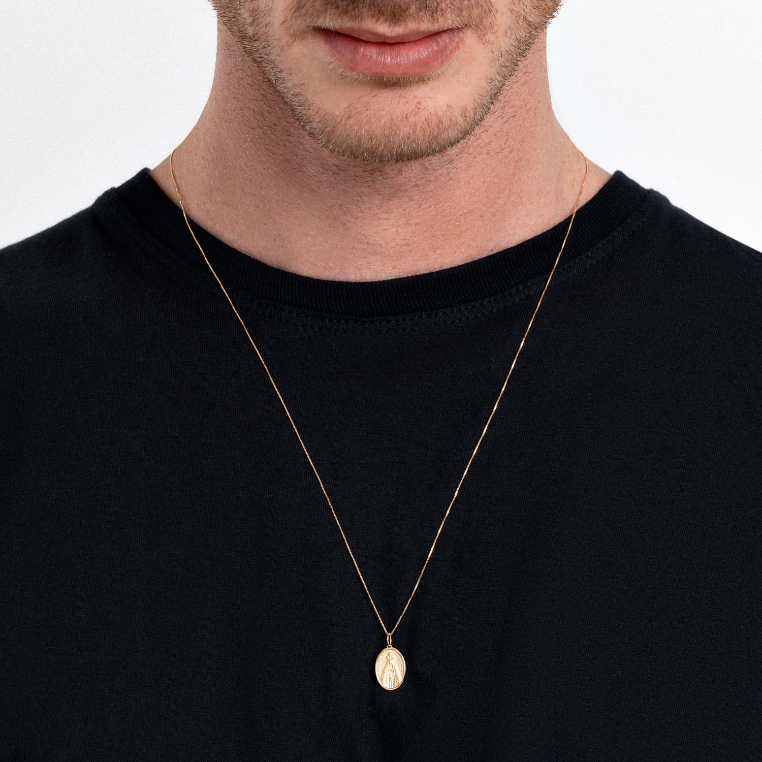 Colar dourado com pingente oval e gravura de uma figura estilizada, usado por um homem com camisa preta.