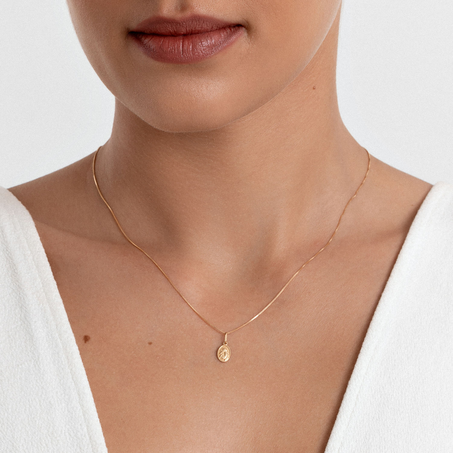 Colar delicado de corrente fina com pingente oval dourado, usado por uma mulher com roupa branca e pele clara.
