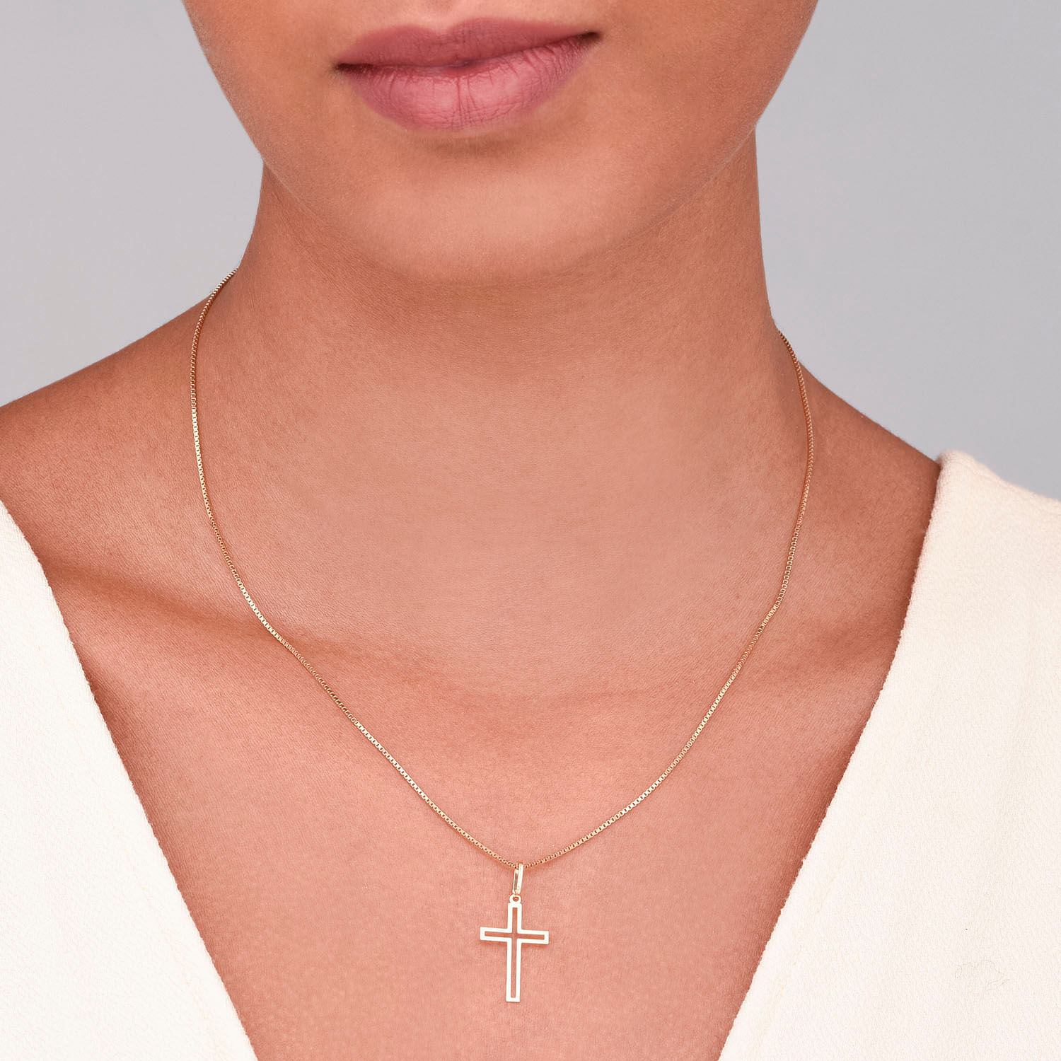Colar com pingente de cruz dourado, usado por uma pessoa com pele clara e camiseta branca.