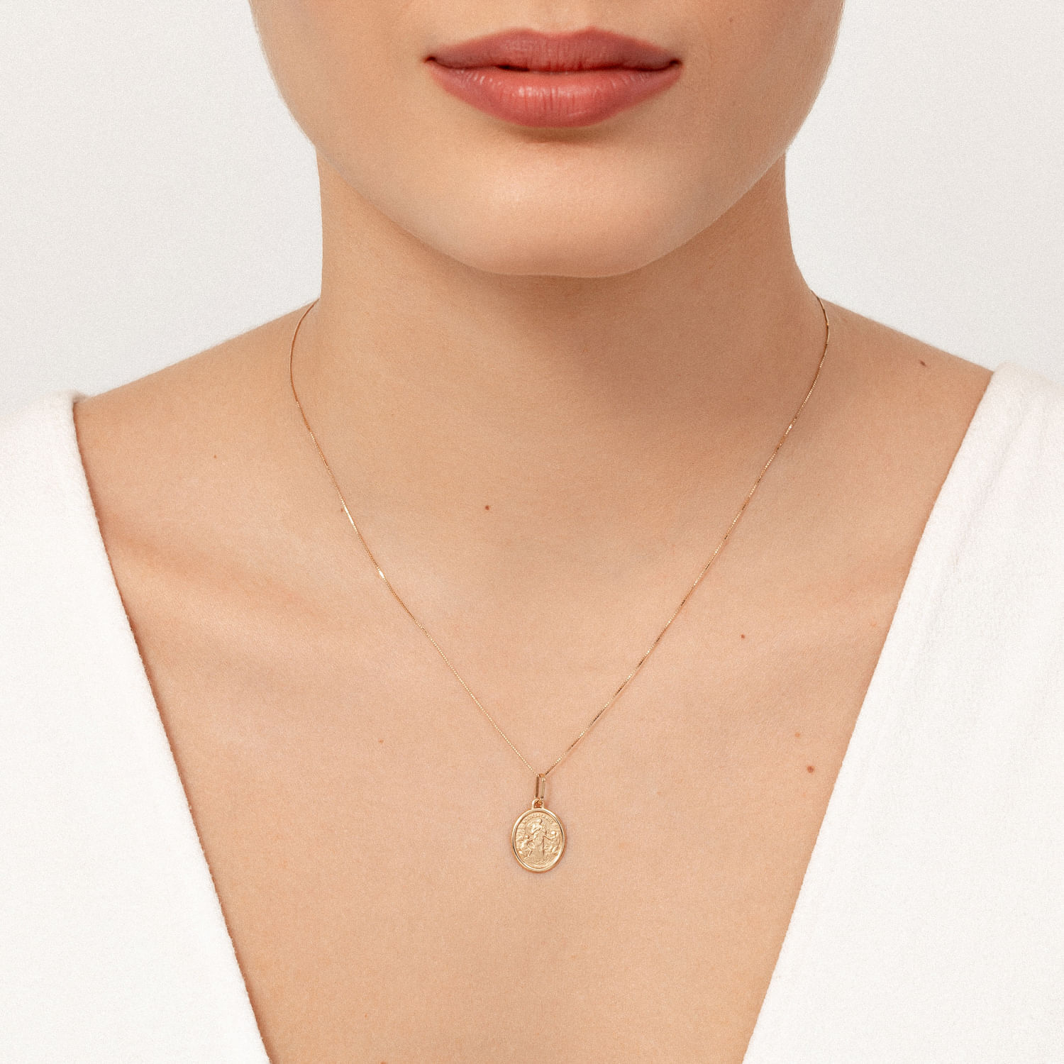 Colar delicado de corrente fina com medalha dourada oval com figura central, usado no pescoço de uma mulher.