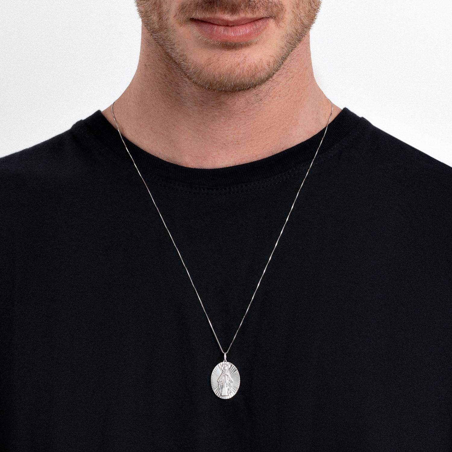 Colar com pingente de medalha religiosa em prata, usado por um homem com camiseta preta.