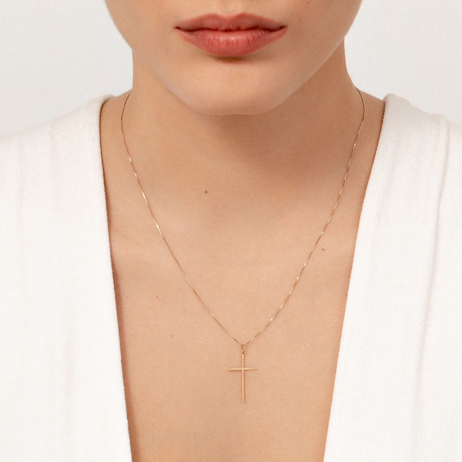 Colar delicado com medalha de cruz dourada, usado por uma pessoa com pele clara e roupas brancas.