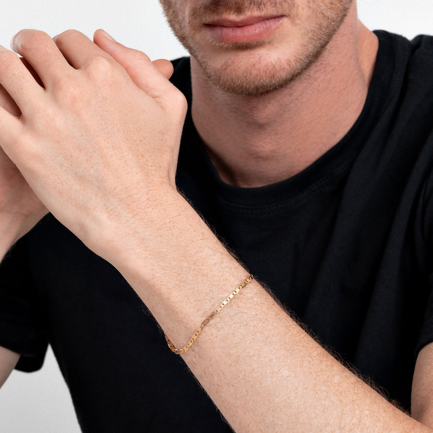Pulso de um homem com pele clara usando uma pulseira fina dourada no braço esquerdo, vestindo uma camiseta preta.