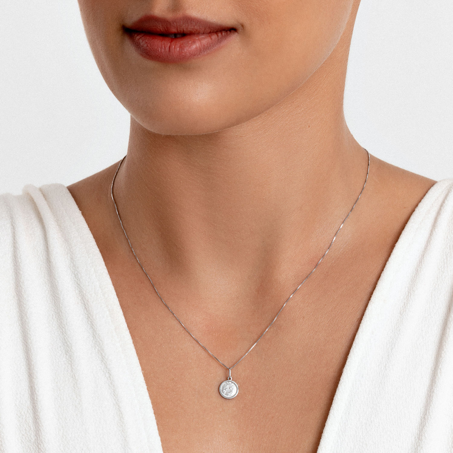 Colar delicado com pingente circular prateado, usada por uma mulher com blusa branca.