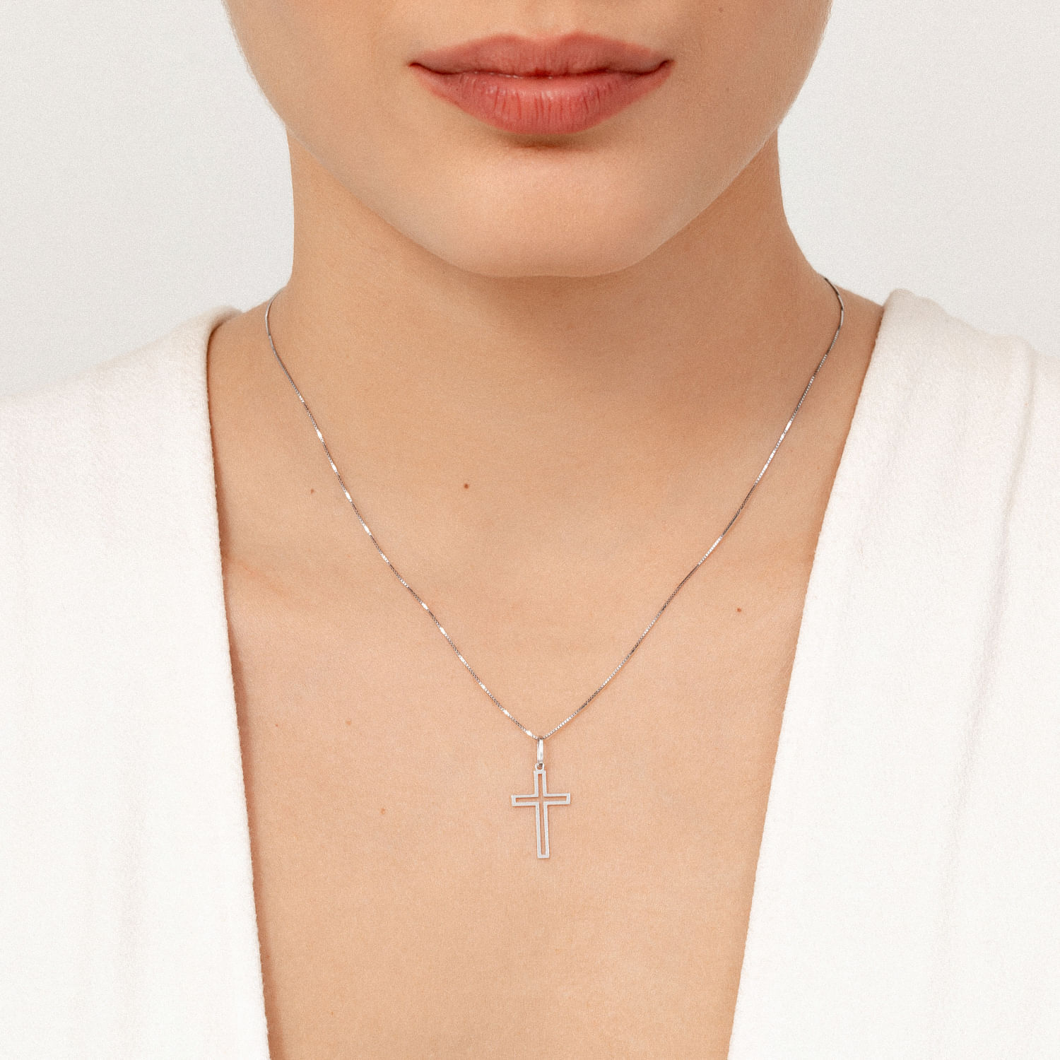 Colar com pingente de cruz prateado, usado por uma mulher com camiseta branca e pele clara.