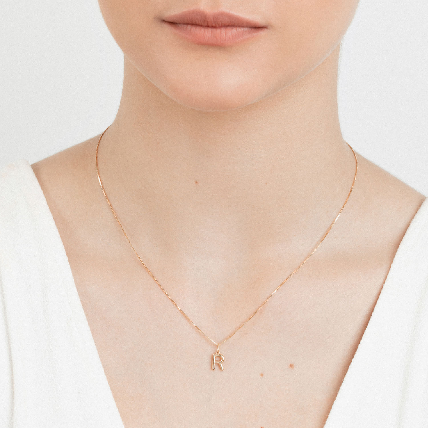 Colar dourado com pingente de letra "R" usado por uma mulher, visível do queixo até o peito.