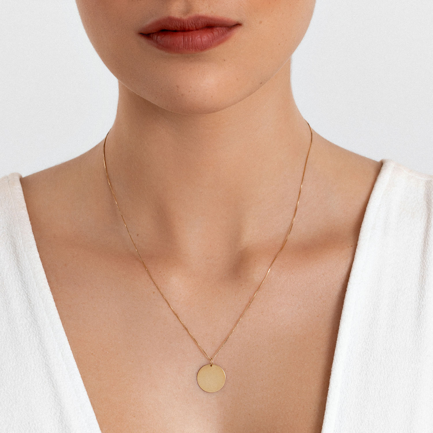 Colar delicado de corrente fina com pingente circular dourado, usado por uma mulher com pele clara e blusa branca.