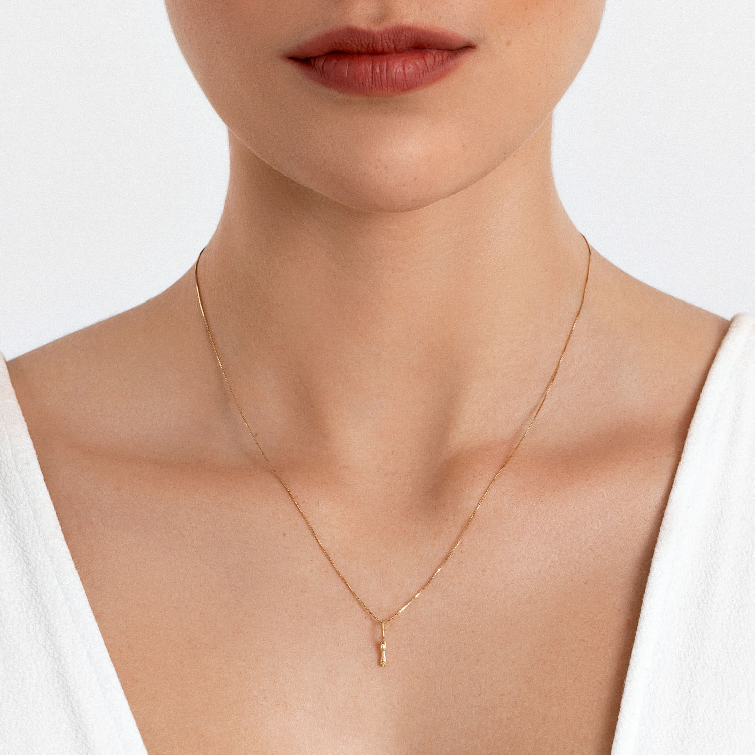 Colar delicado de corrente dourada com pingente pequeno, usado por uma pessoa com pele clara, veste uma blusa branca.