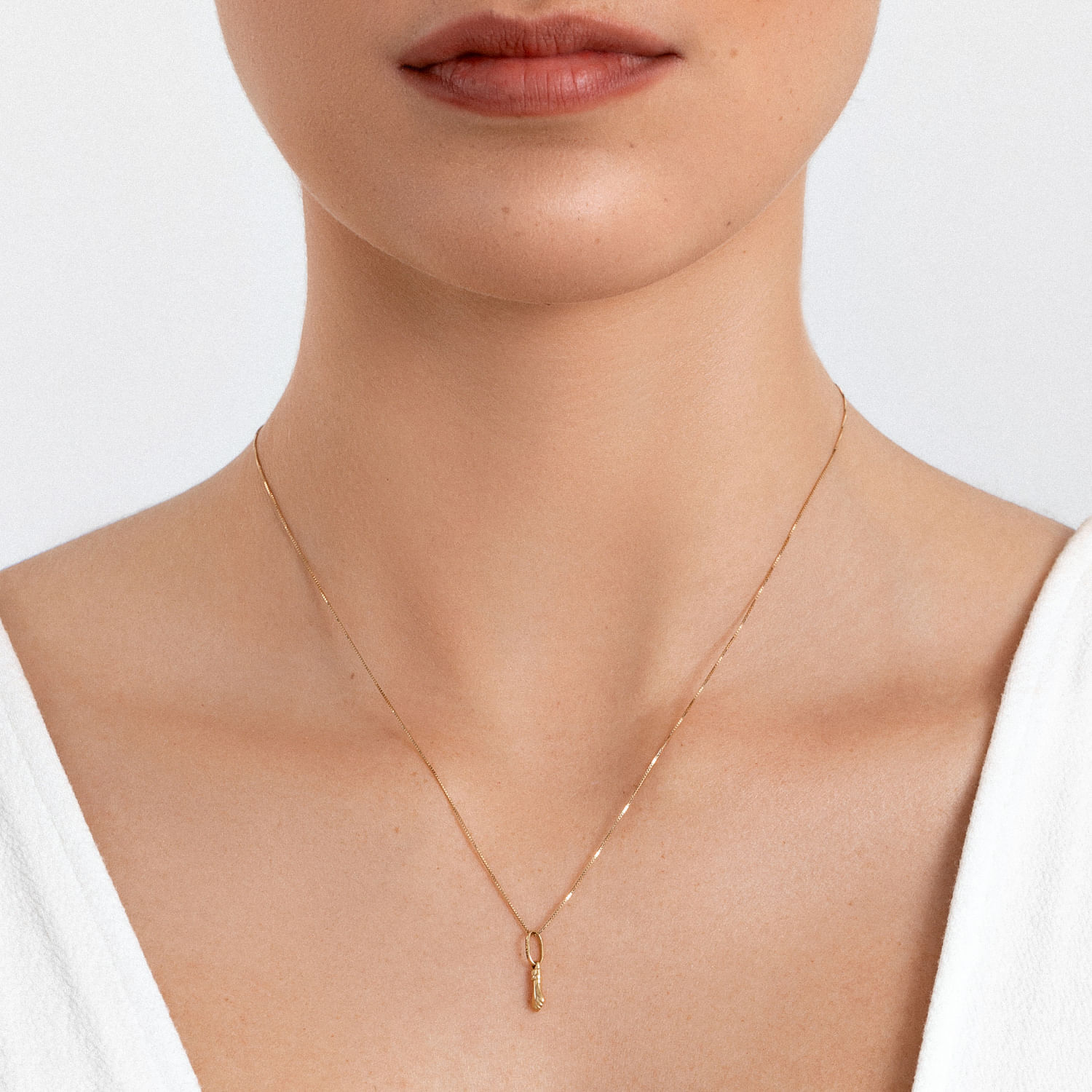 Colar dourado delicado com pingente em forma de lágrima, usado por uma pessoa com pele clara e camisa branca.