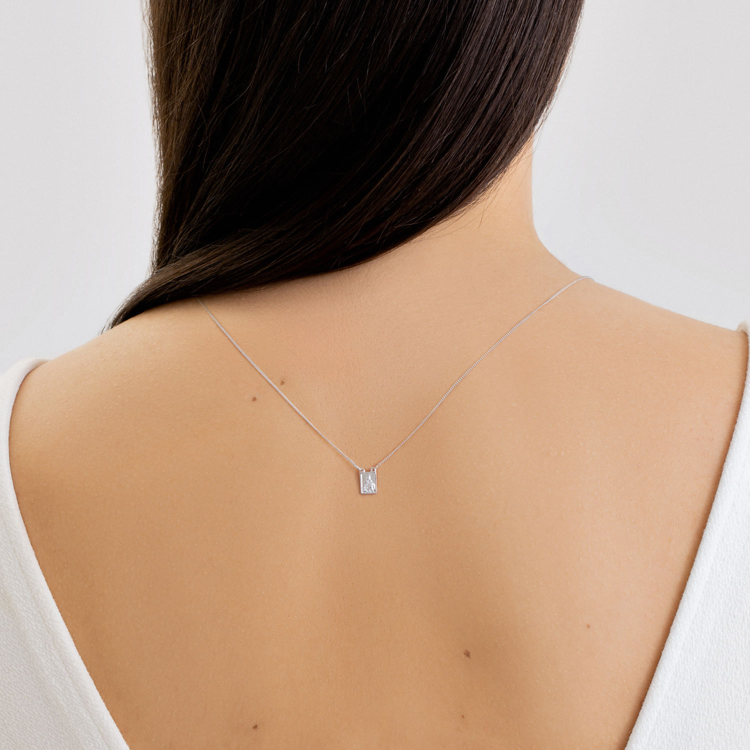 Colar delicado com pingente retangular, usado na parte de trás do pescoço, sobre pele clara, com cabelo escuro e vestido branco.