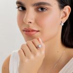 Mulher com pele clara e olhos claros, usando brinco e anel com diamantes, posando com a mão próxima ao rosto e usando uma corrente fina.
