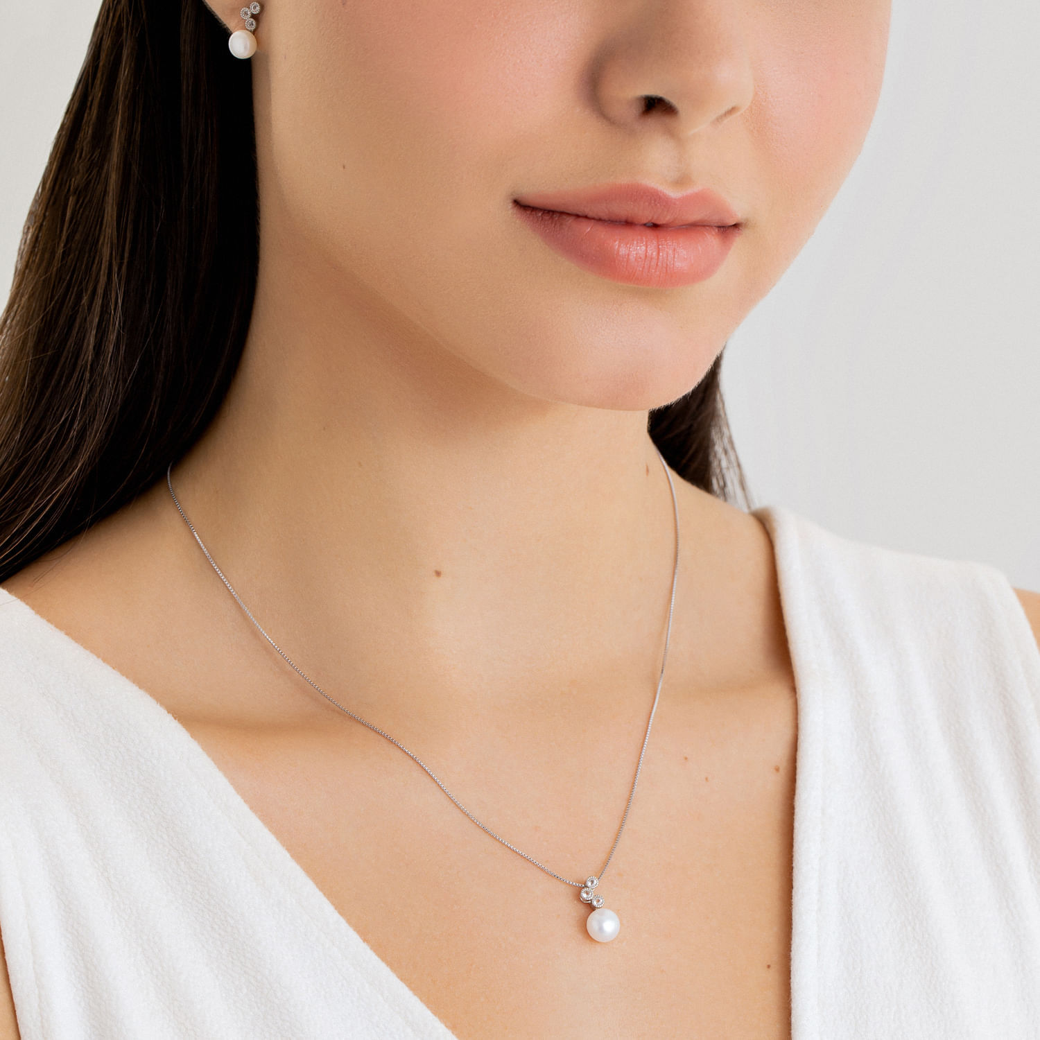 Colar com pingente de pérola e detalhes brilhantes, usado por uma mulher de pele clara e cabelos lisos escuros.