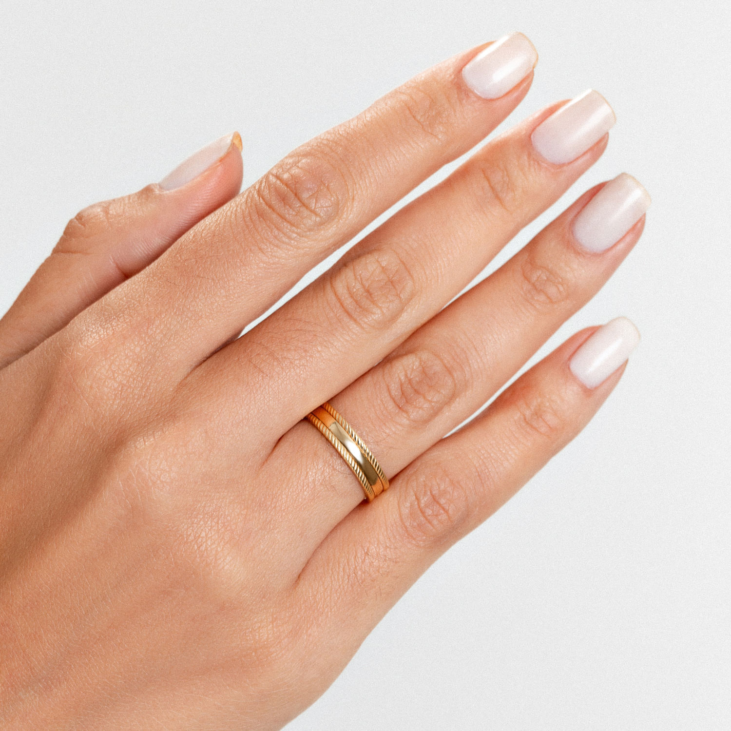 Mão com unhas claras e bem feitas usando duas alianças douradas, contra fundo branco.
