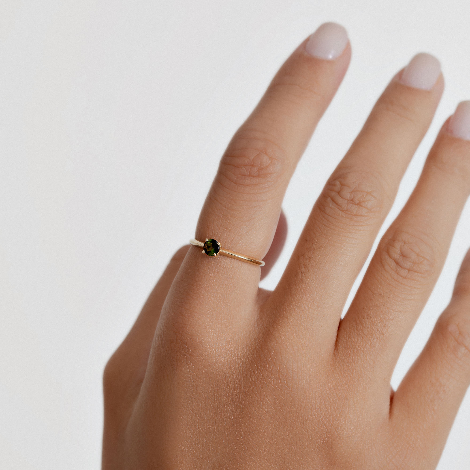 Anel delicado com pedra verde em uma mão com unhas naturais, contra fundo branco.