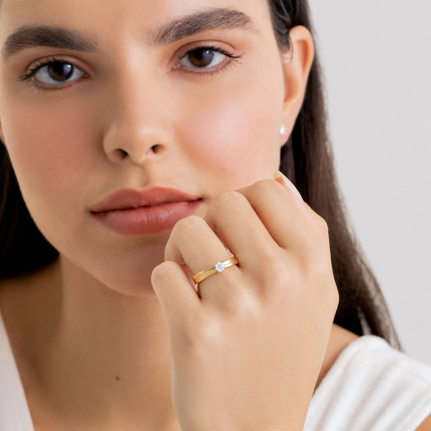 Close-up de uma mulher com pele clara e cabelos castanhos, usando um anel dourado com uma pedra brilhante na mão esquerda, apoiada no queixo.