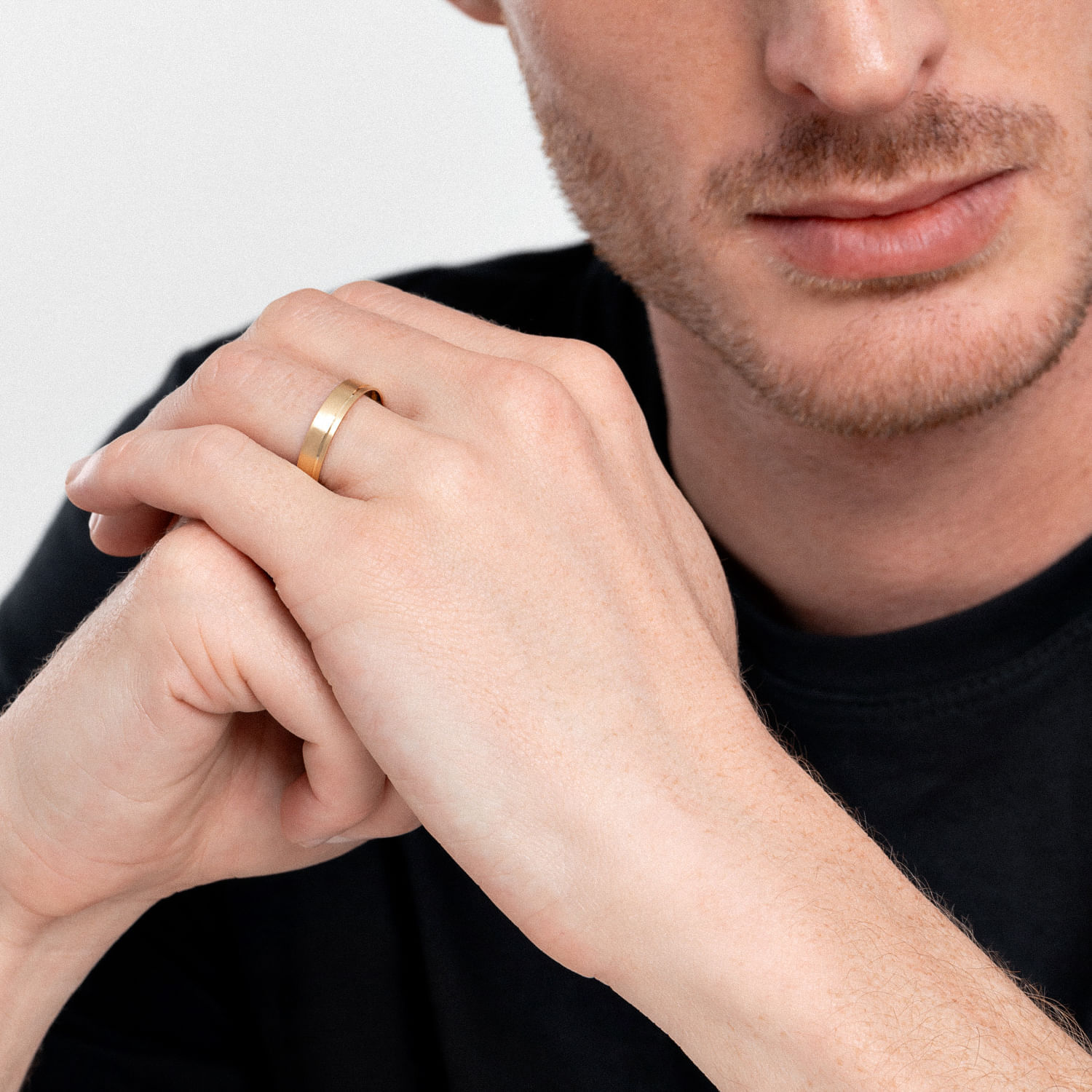 Mão de homem com barba, usando anel dourado na mão esquerda, apoiada no queixo, vestindo camiseta preta.