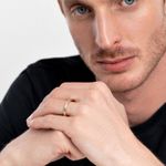 Homem com cabelo castanho claro, olhos azuis, usando uma camiseta preta, exibe uma mão com uma aliança dourada no dedo indicador.