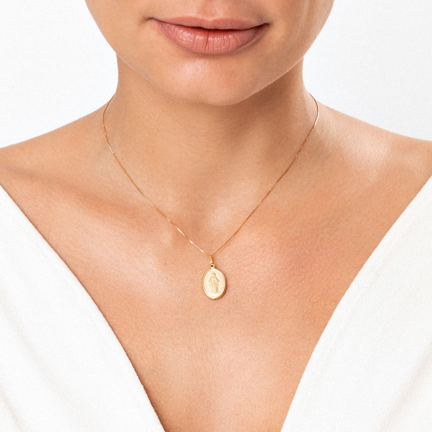 Colar de corrente fina dourada com pingente ovalino contendo uma figura religiosa, usado por uma mulher com pele clara e vestido branco.