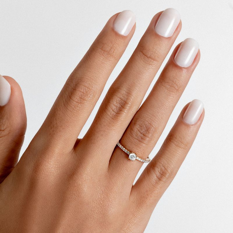 Dedo com unhas bem cuidadas usando esmalte branco e anel de noivado com uma pedra central grande e pequenas pedras ao redor.