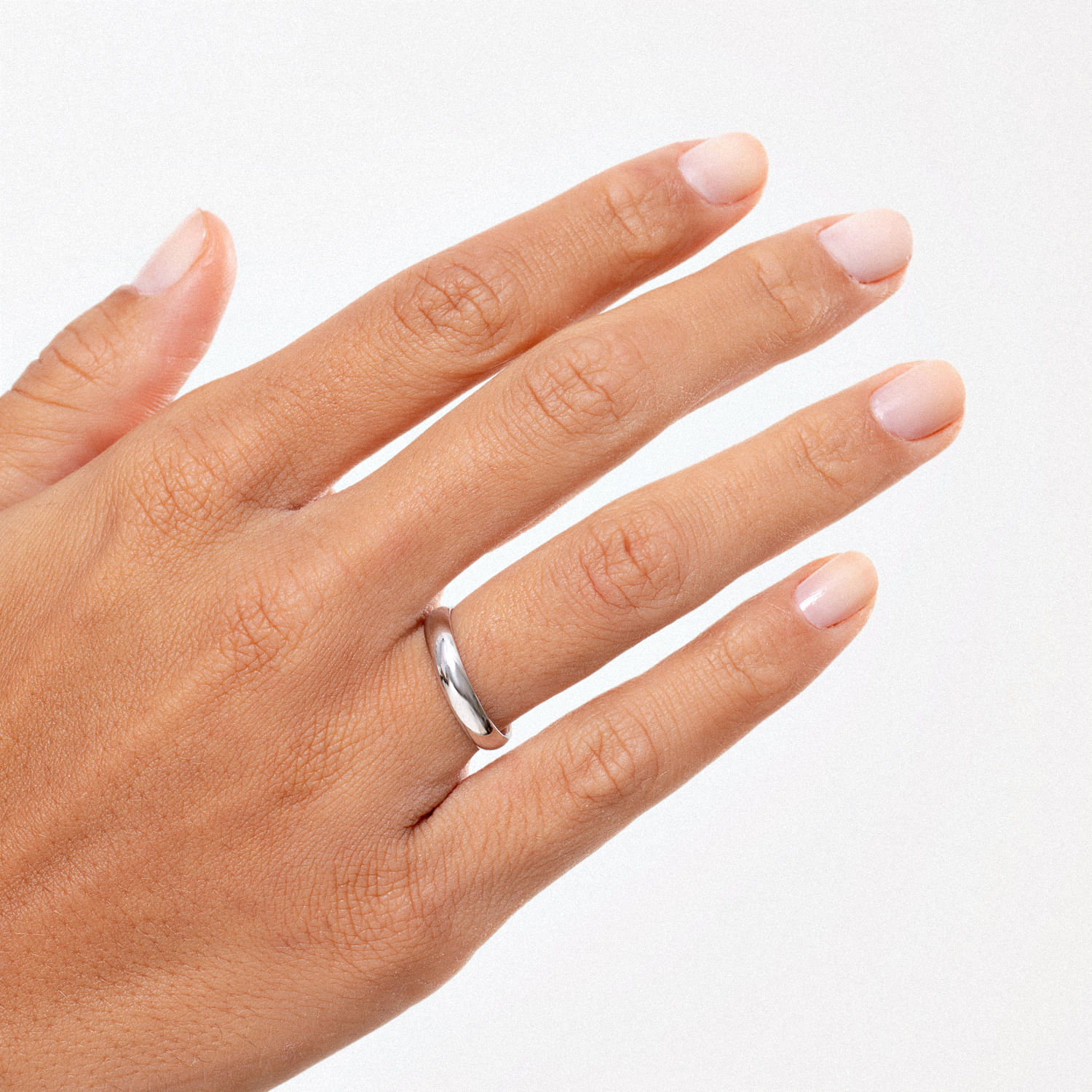 Mão com unhas pintadas de nude, usando aliança de prata na quarta dedo. Fundo branco claro.