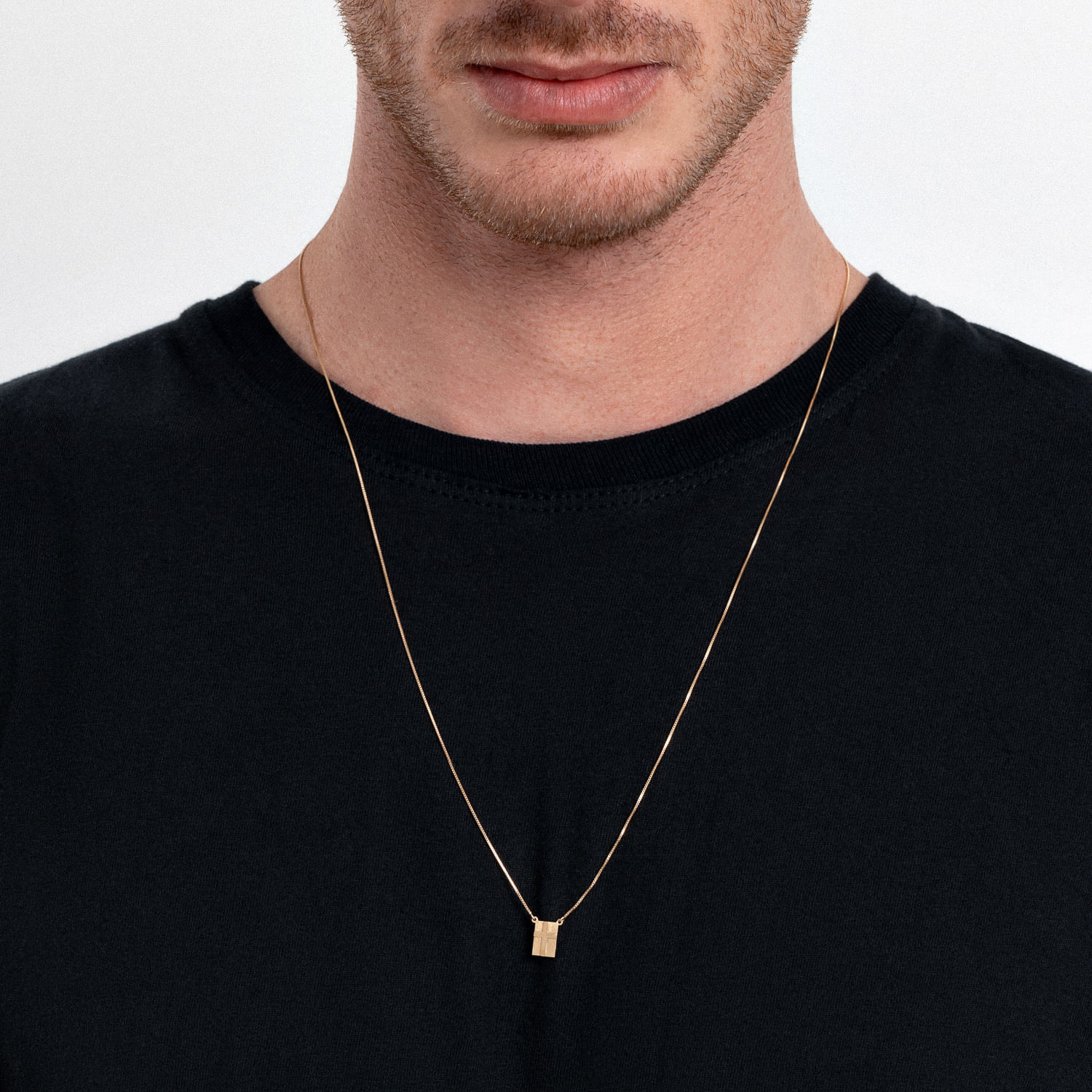 Colar dourado com pingente retangular, usado por um homem com camiseta preta.