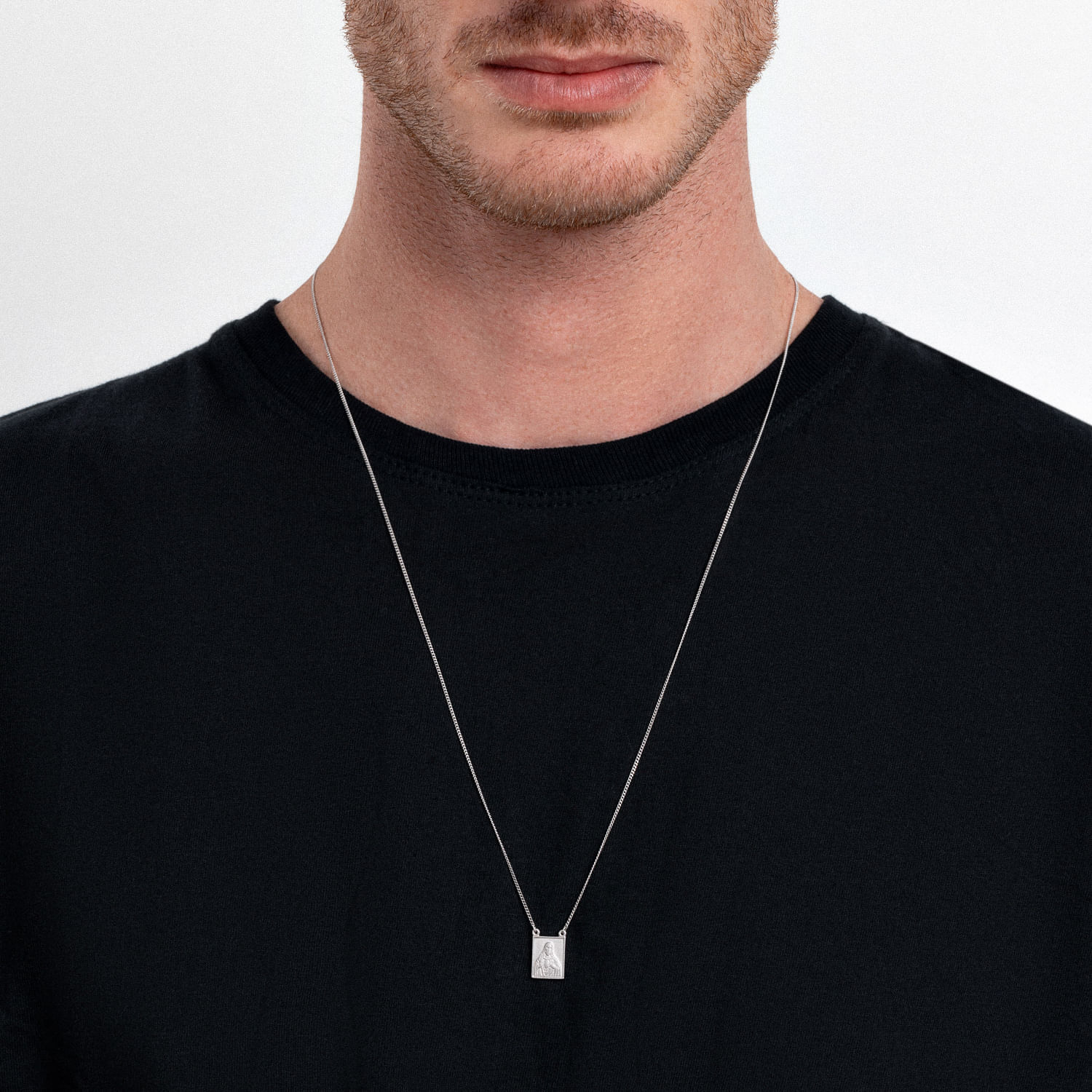 Homem usando uma camiseta preta e um colar com pingente retangular prateado, visível na imagem.