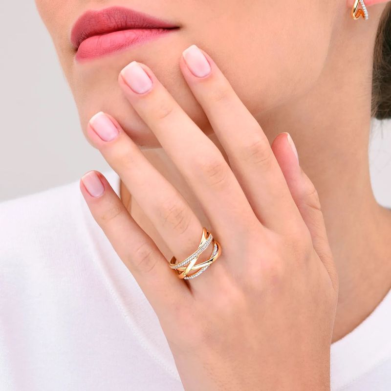 Dedo de uma mulher com unhas bem cuidadas usando um anel dourado com detalhes prateados, perto do rosto com batom cor de rosa.