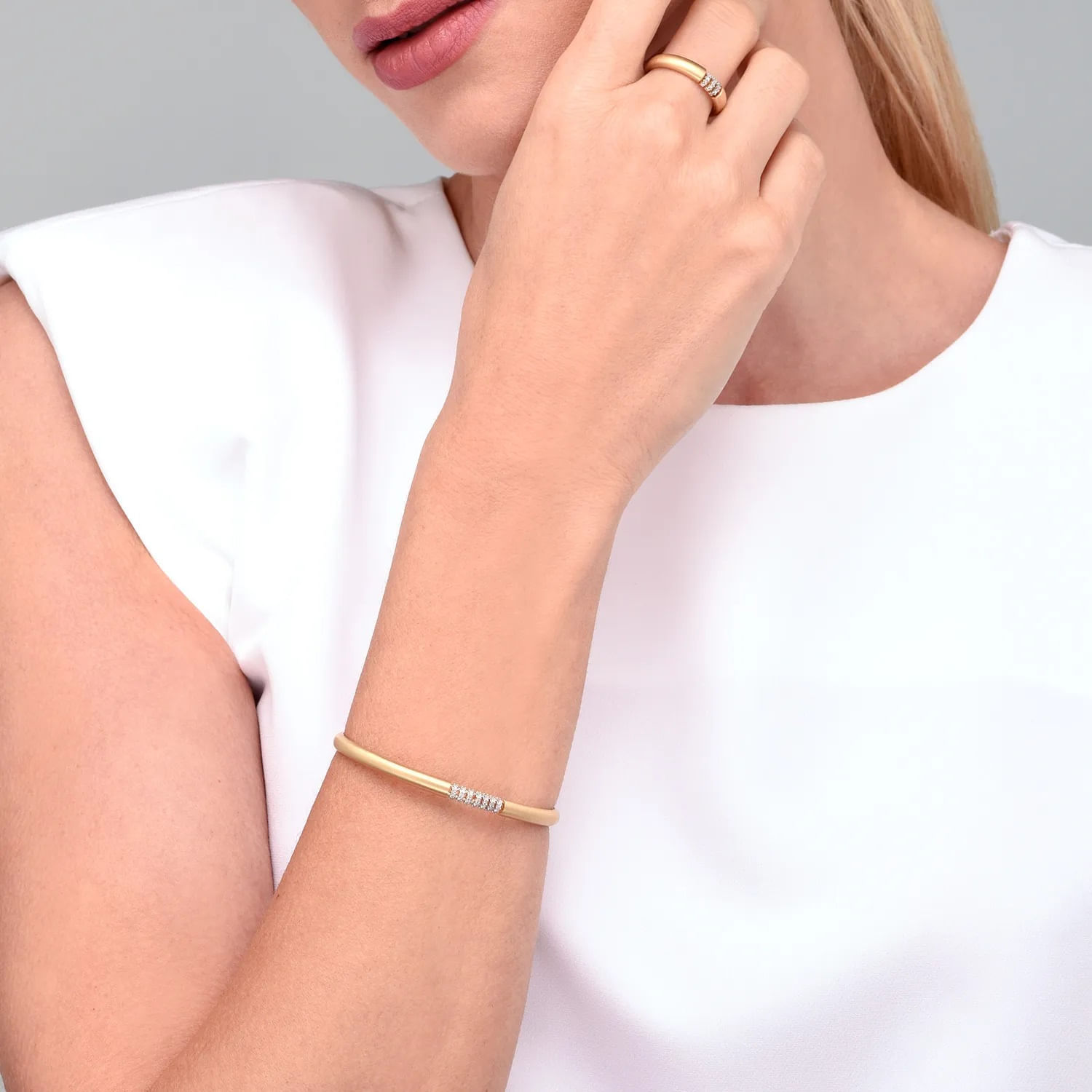 Close-up de uma mulher usando uma pulseira e um anel dourados, com pele clara, vestindo blusa branca e cabelo loiro.