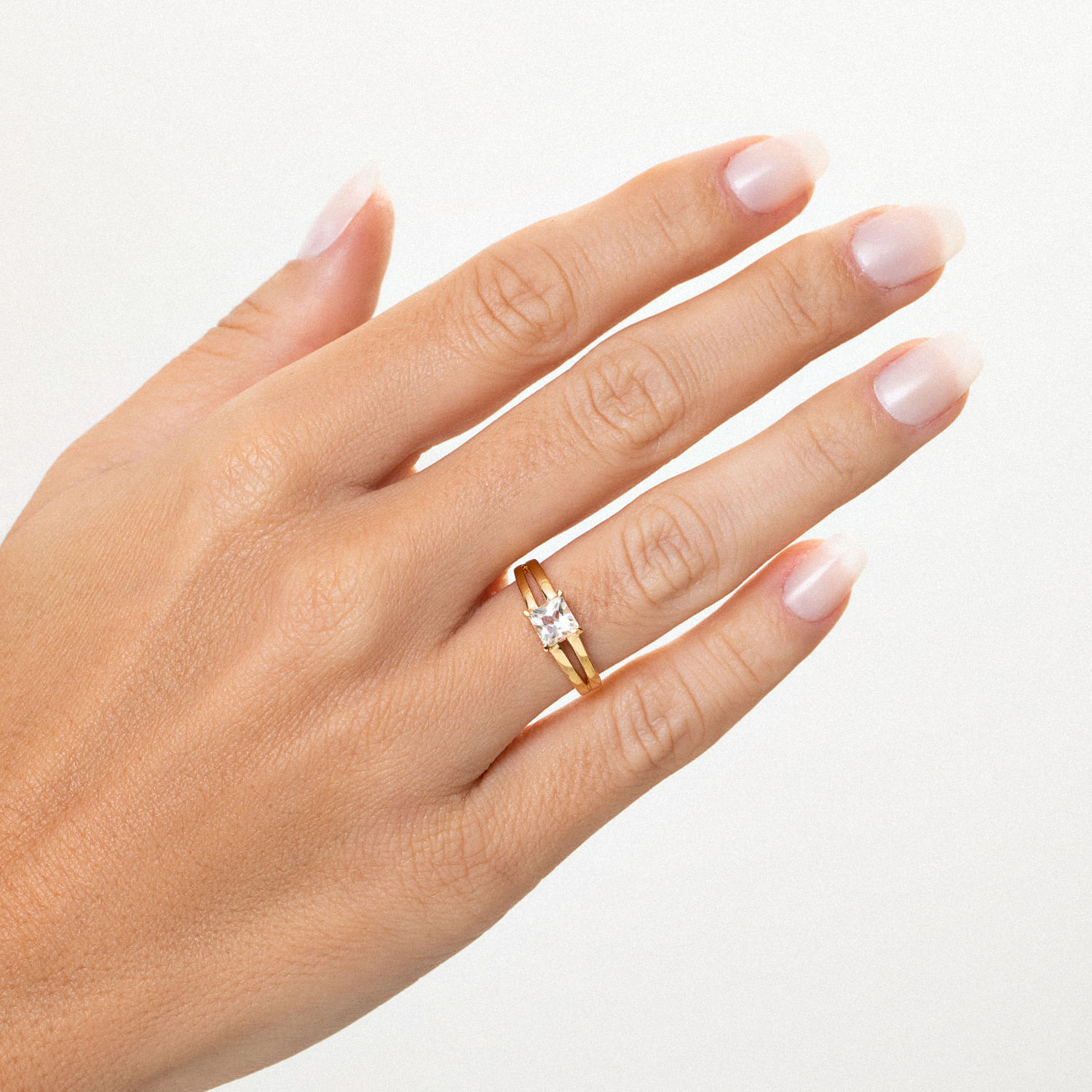 Mão com pele clara, exibindo um anel dourado com uma pedra transparente no anelar. Fundo branco.