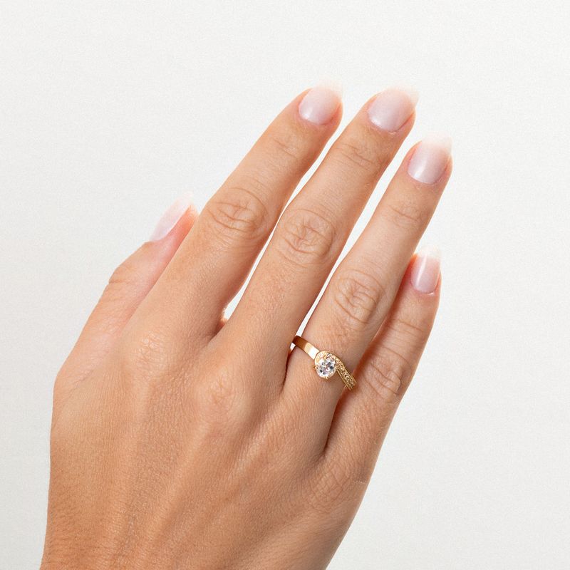 Dedo com anel de ouro com uma pedra brilhante arredondada no centro, em fundo claro.