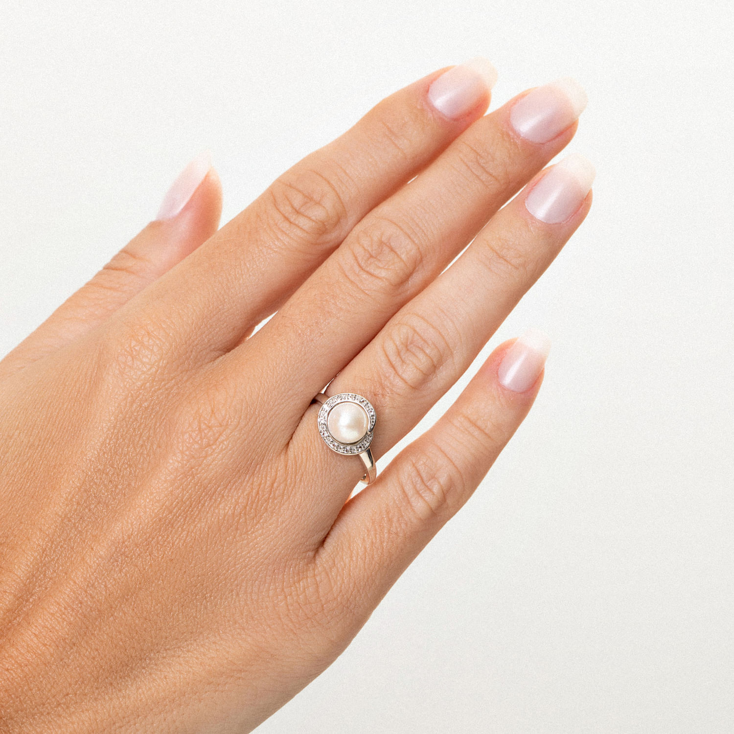 Anel com pérola central cercada por pequenos diamantes, usado na dedo anelar de uma mão com unhas bem cuidadas.